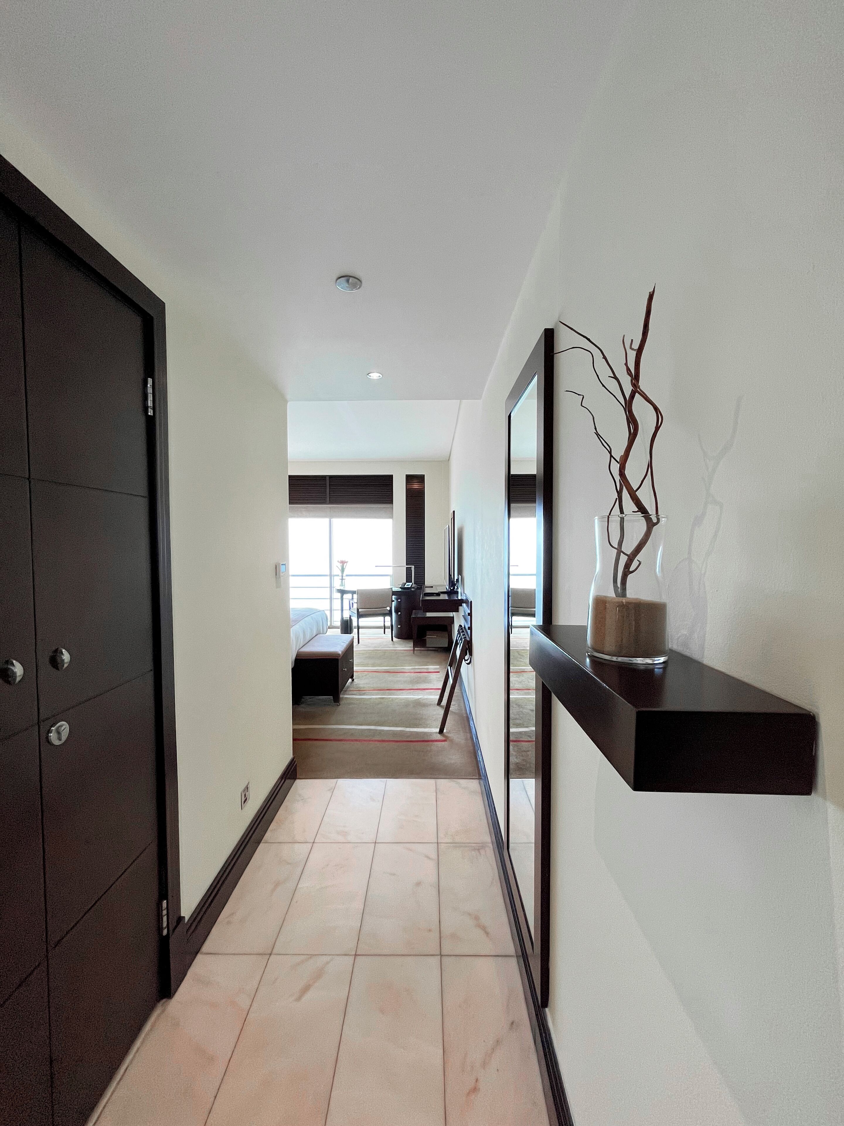 A modern hotel hallway with a dark wooden door, a glass vase with branches, and a bed in the background.