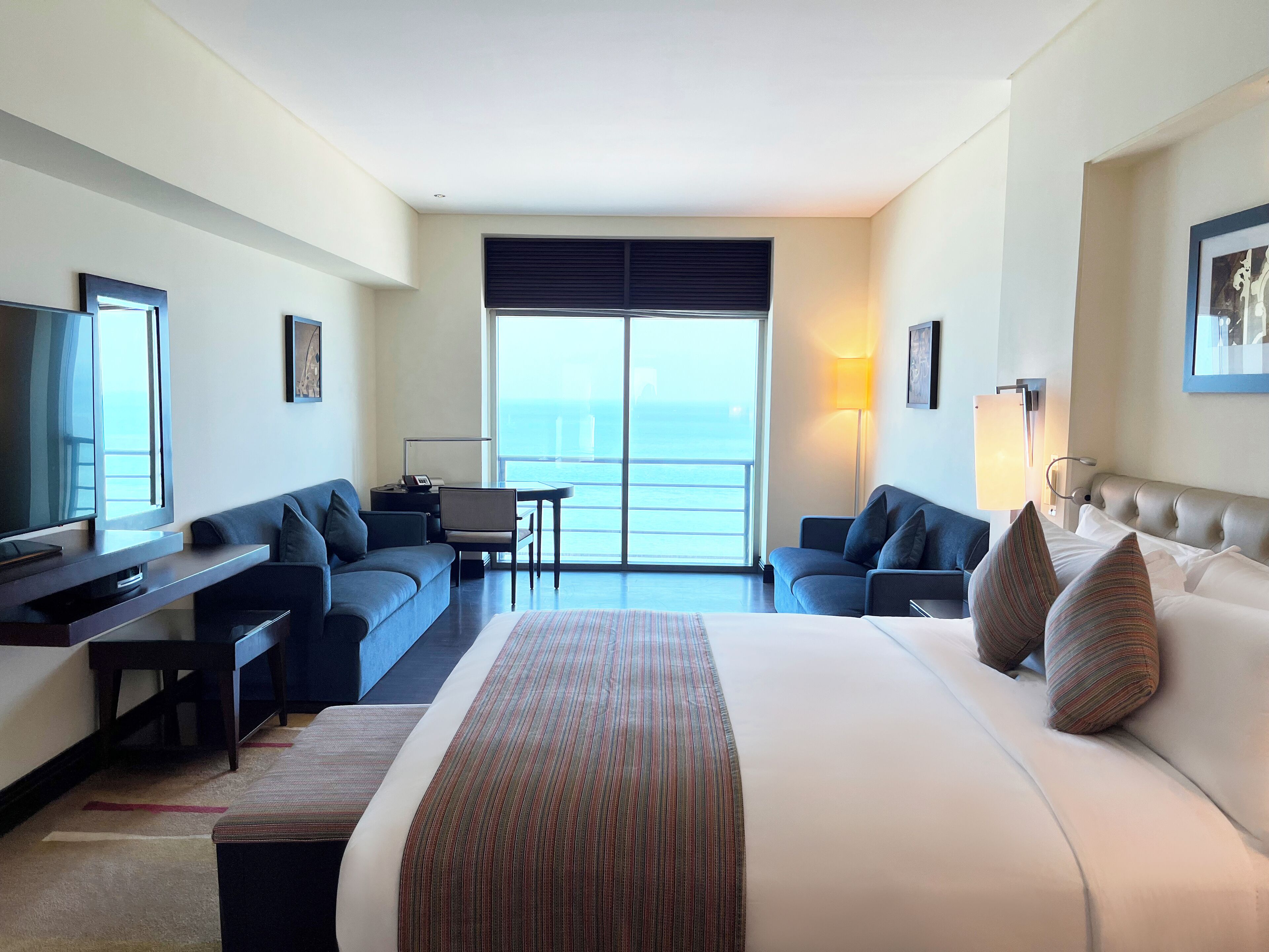 Ein Hotelzimmer mit einem großen Bett, einem Schreibtisch, einer Couch und einem Fernseher. Es gibt einen Balkon mit Blick aufs Meer.