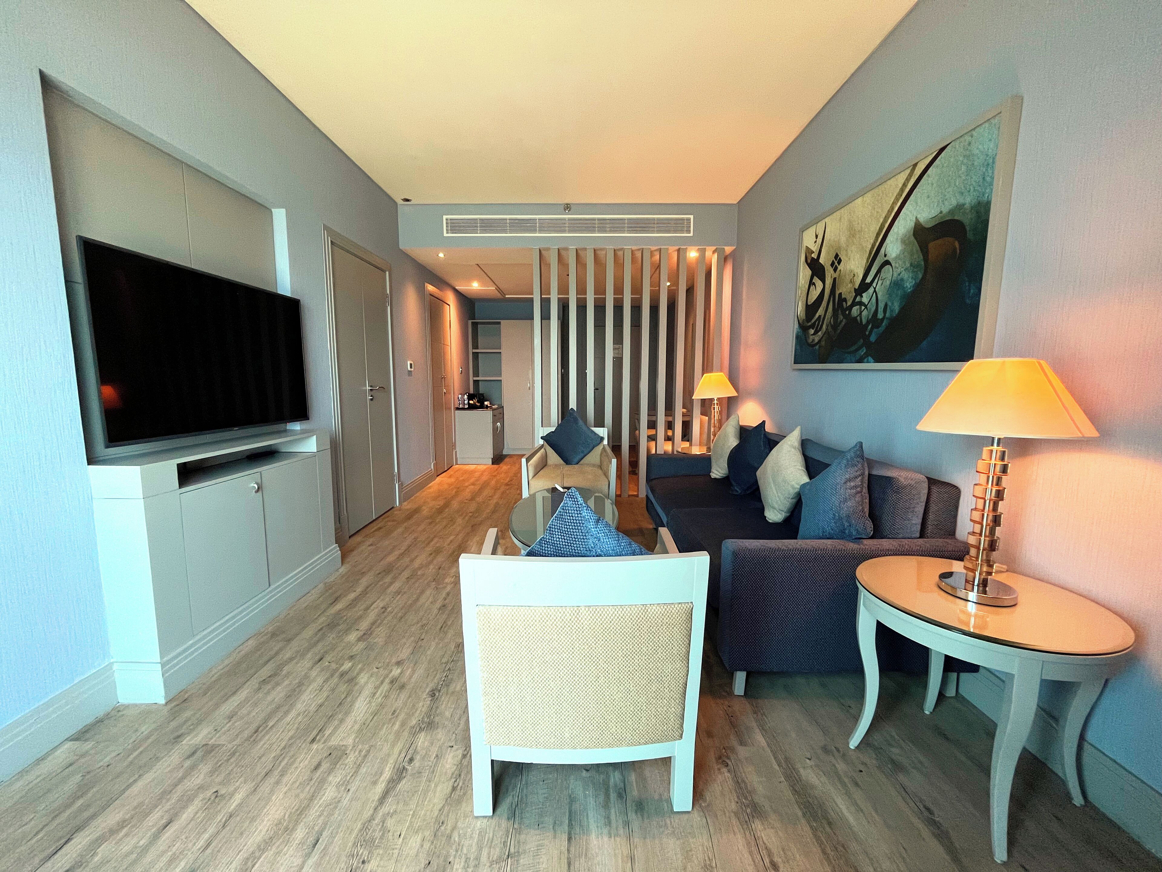 A modern hotel room with a flat-screen TV, a sofa, a chair, a small table, and a lamp.