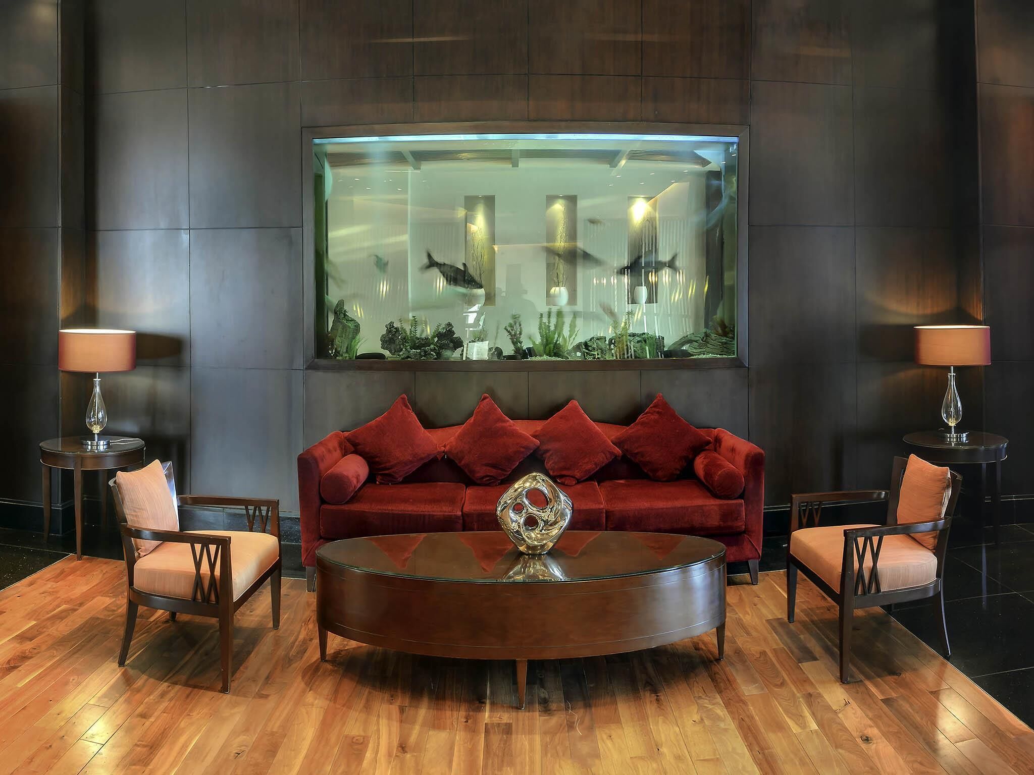 A modern living room with a red velvet sofa, two armchairs, a glass coffee table, and a large aquarium above the sofa.