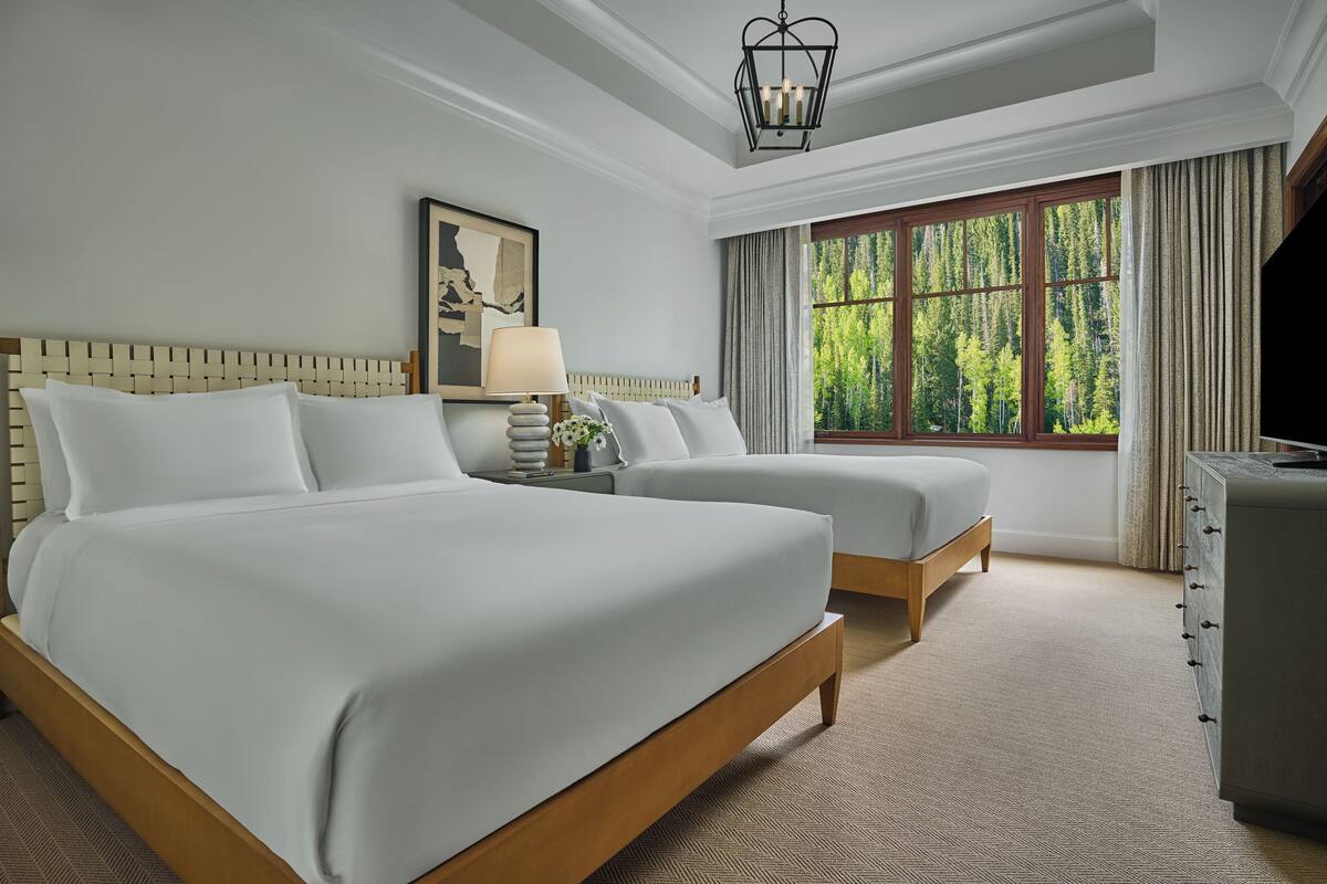 Une chambre d’hôtel avec deux lits, une table de chevet avec une lampe, un tableau, une fenêtre avec des rideaux et une commode.