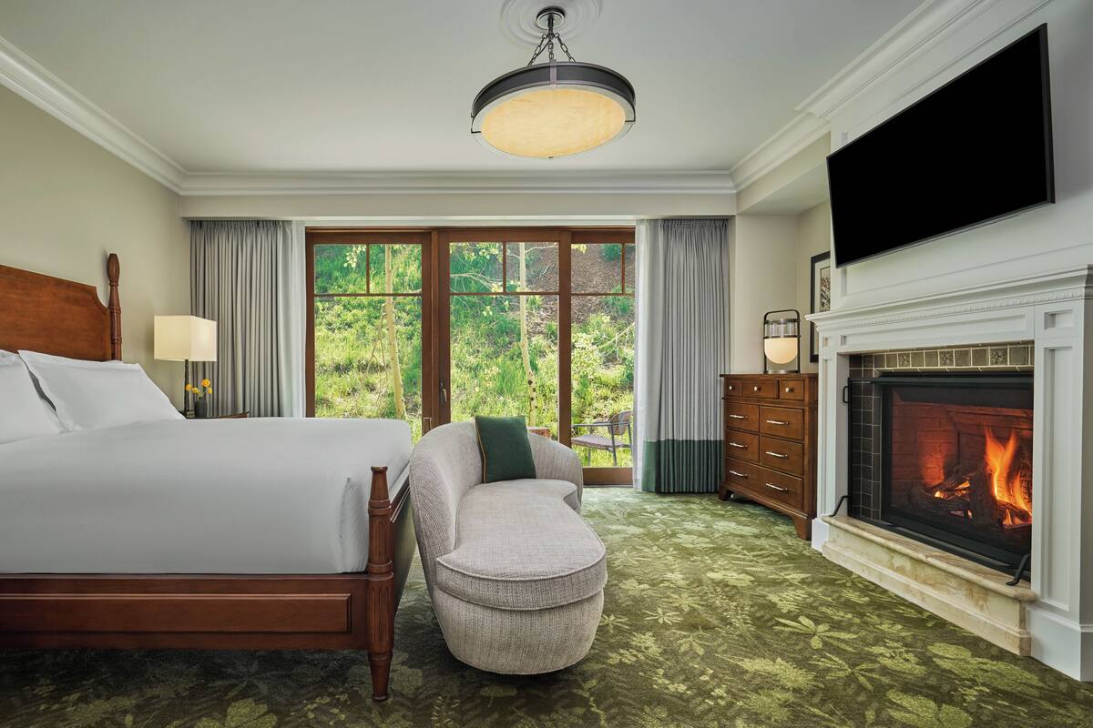 Une chambre d’hôtel comprenant un lit, un coin salon, une cheminée et une vue sur la verdure par les fenêtres.