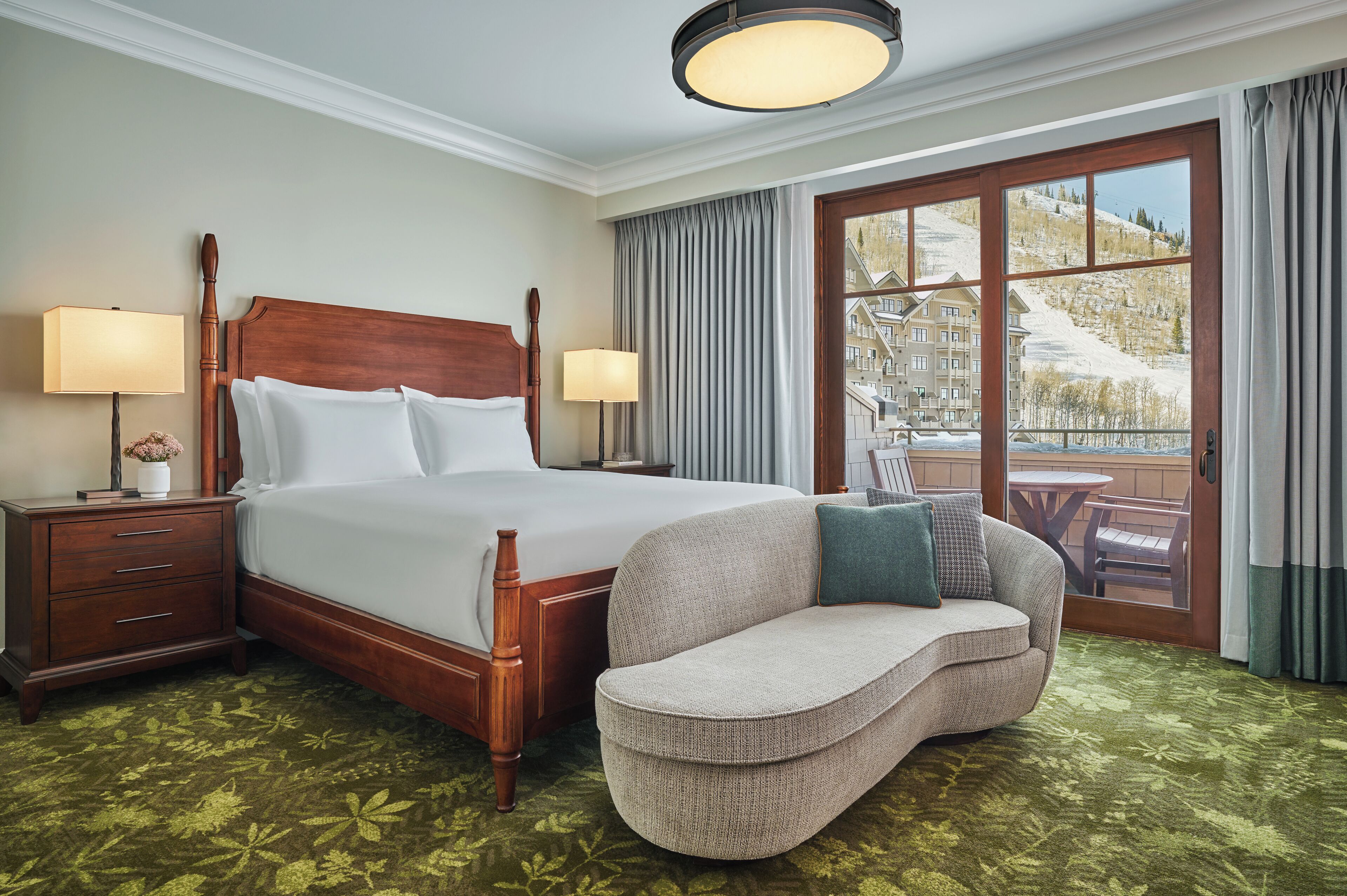 Quarto de hotel com uma cama grande, duas luminárias de cabeceira, uma mesinha de cabeceira de madeira, um tapete verde estampado, um sofá e vista para uma montanha nevada através de uma grande janela.