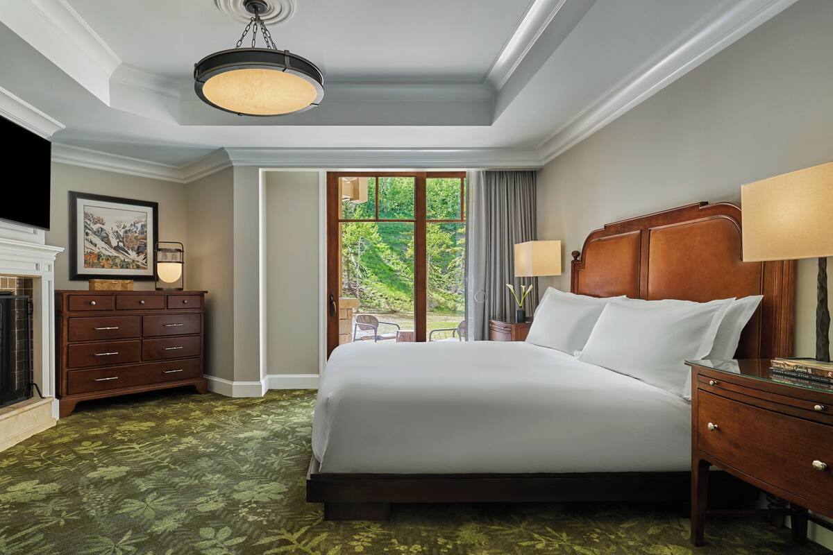Une chambre d’hôtel avec une cheminée, un grand lit, une table de chevet, une commode et une vue sur l’extérieur.