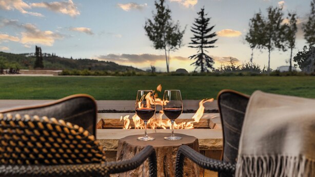 A cozy outdoor seating area with wicker chairs and a round table, featuring a fire pit and two glasses of red wine.