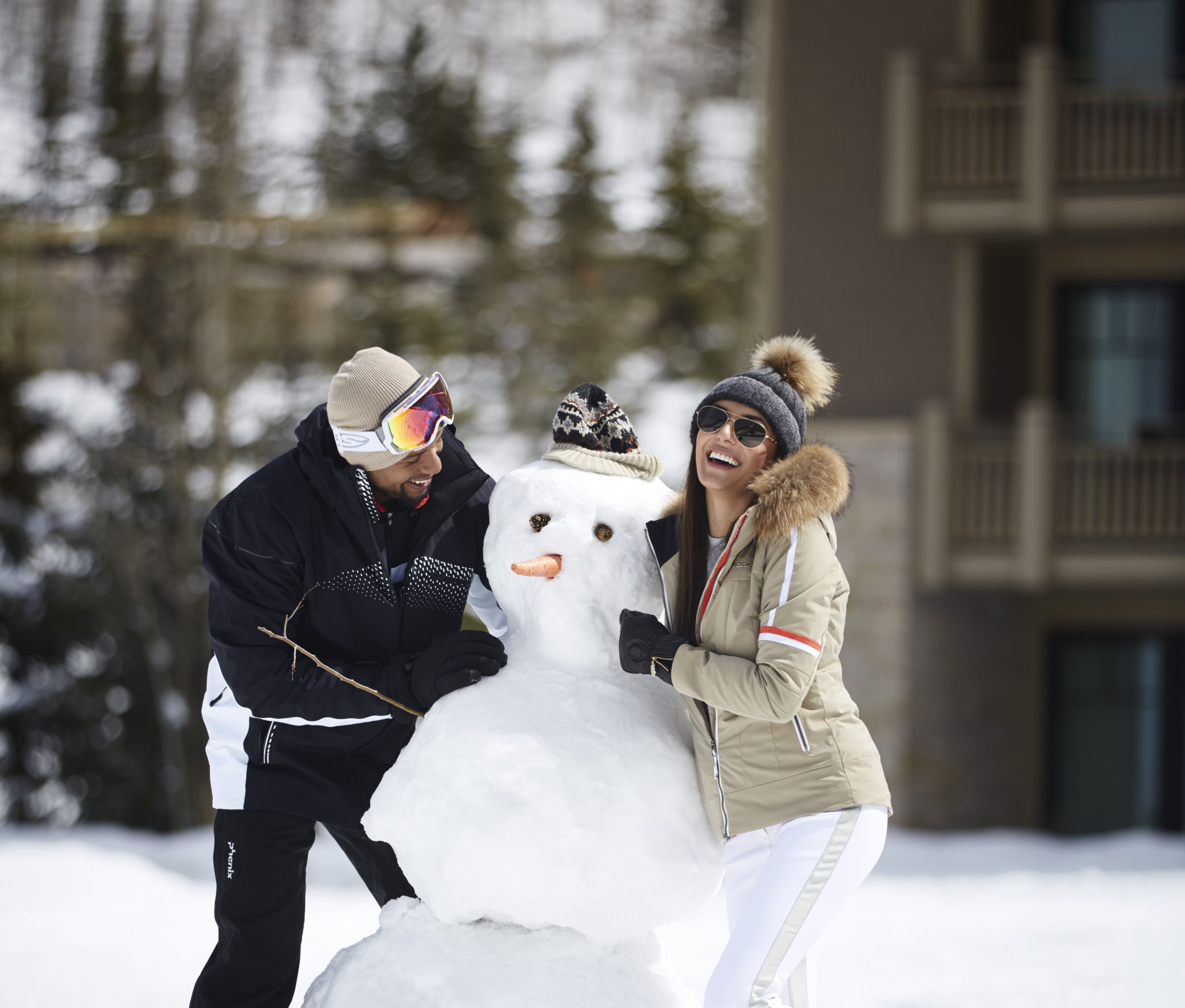 A snowman being built by two people in a snowy outdoor area with a building in the background.