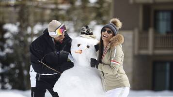 A snowman being built by two people in a snowy outdoor area with a building in the background.