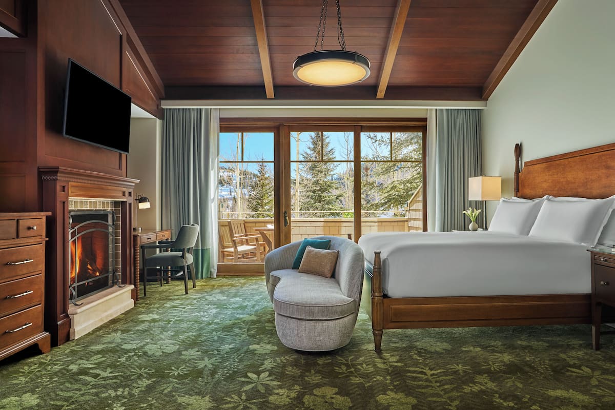 Une chambre d’hôtel avec un lit, un bureau, une chaise, une cheminée et une vue sur les arbres, visible à travers de grandes fenêtres.