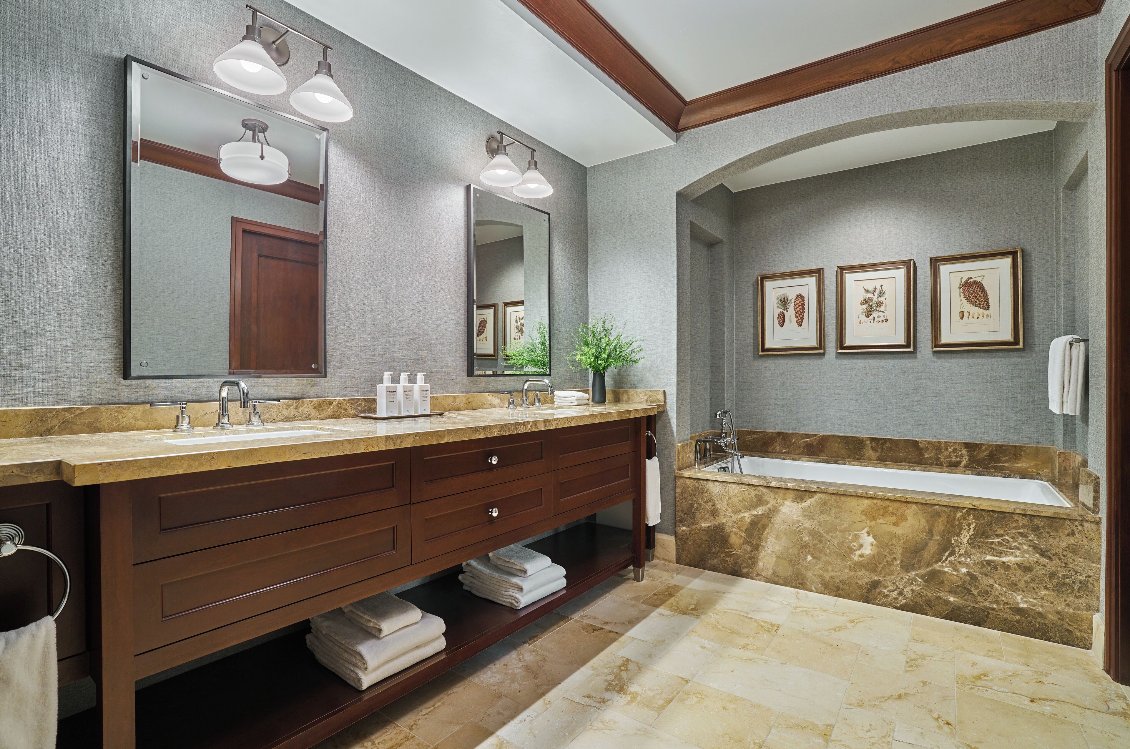 A bathroom with a double vanity, a large mirror, two wall-mounted lights, a bathtub, and framed botanical prints on the wall.
