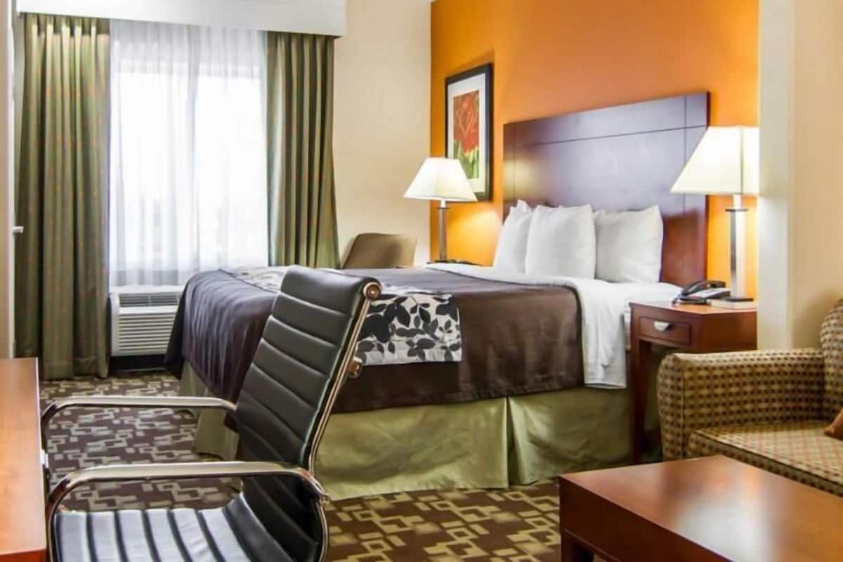 A hotel room with a bed, two bedside lamps, a chair, a desk, and a window with curtains.