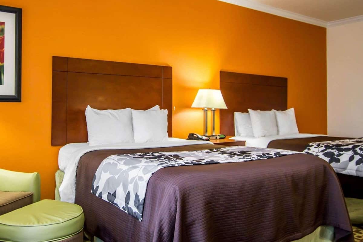 A hotel room with two beds, a nightstand with a lamp, and a green armchair.