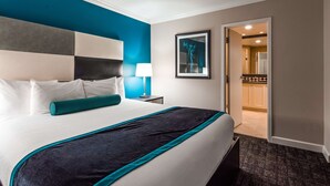 Een hotelkamer met een groot bed, een nachtkastje met een lamp, een ingelijst schilderij aan de muur, en een open deur naar een badkamer.