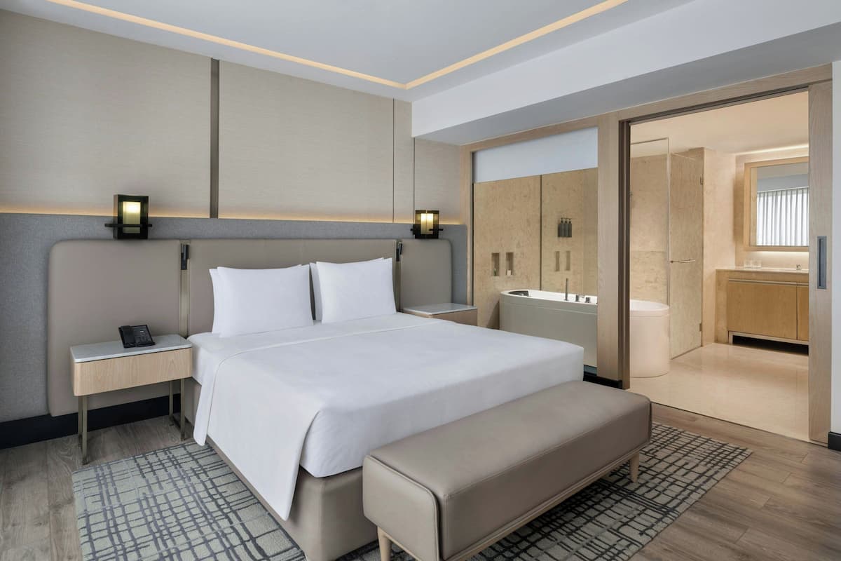 A modern hotel room with a large bed, bedside tables, a bench, a walk-in closet, and a bathroom with a mirror and sink.