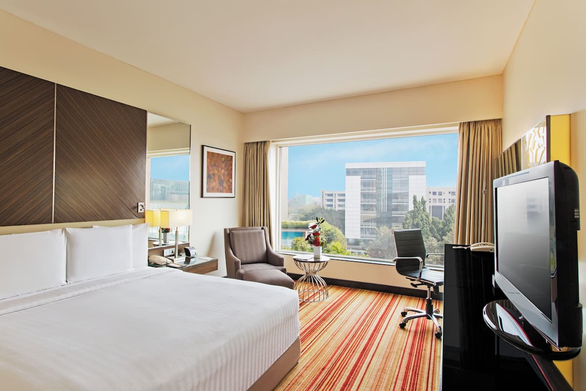 A hotel room with a large bed, a TV, a desk with a lamp, a chair, and a view of the city.