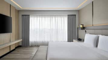Una habitación de hotel moderna con una cama grande, un televisor de pantalla plana montado en la pared, una mesita de noche con lámpara y un amplio ventanal con cortinas.