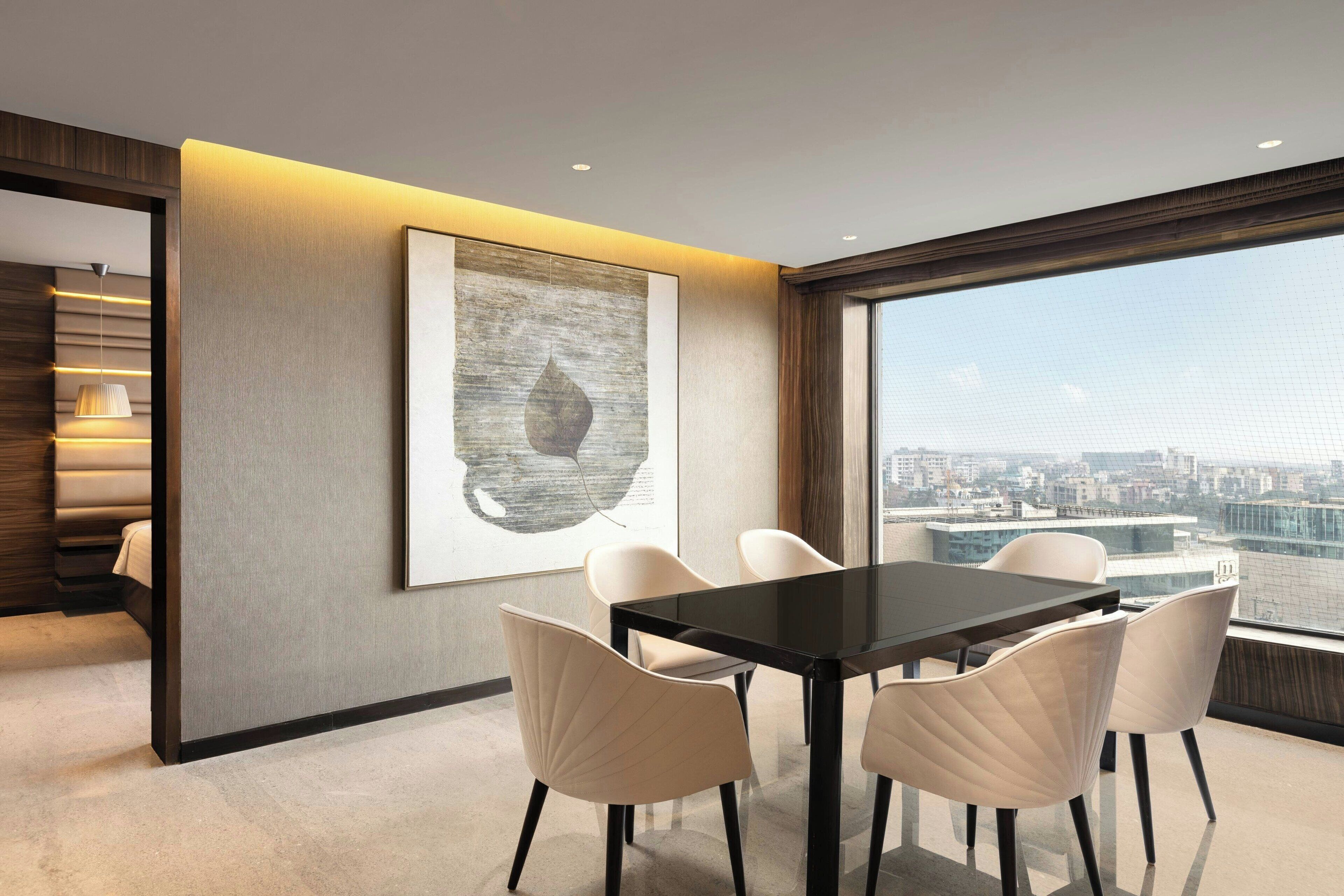 A modern dining area with a black table and white chairs, a large abstract painting on the wall, and a city view through large windows.
