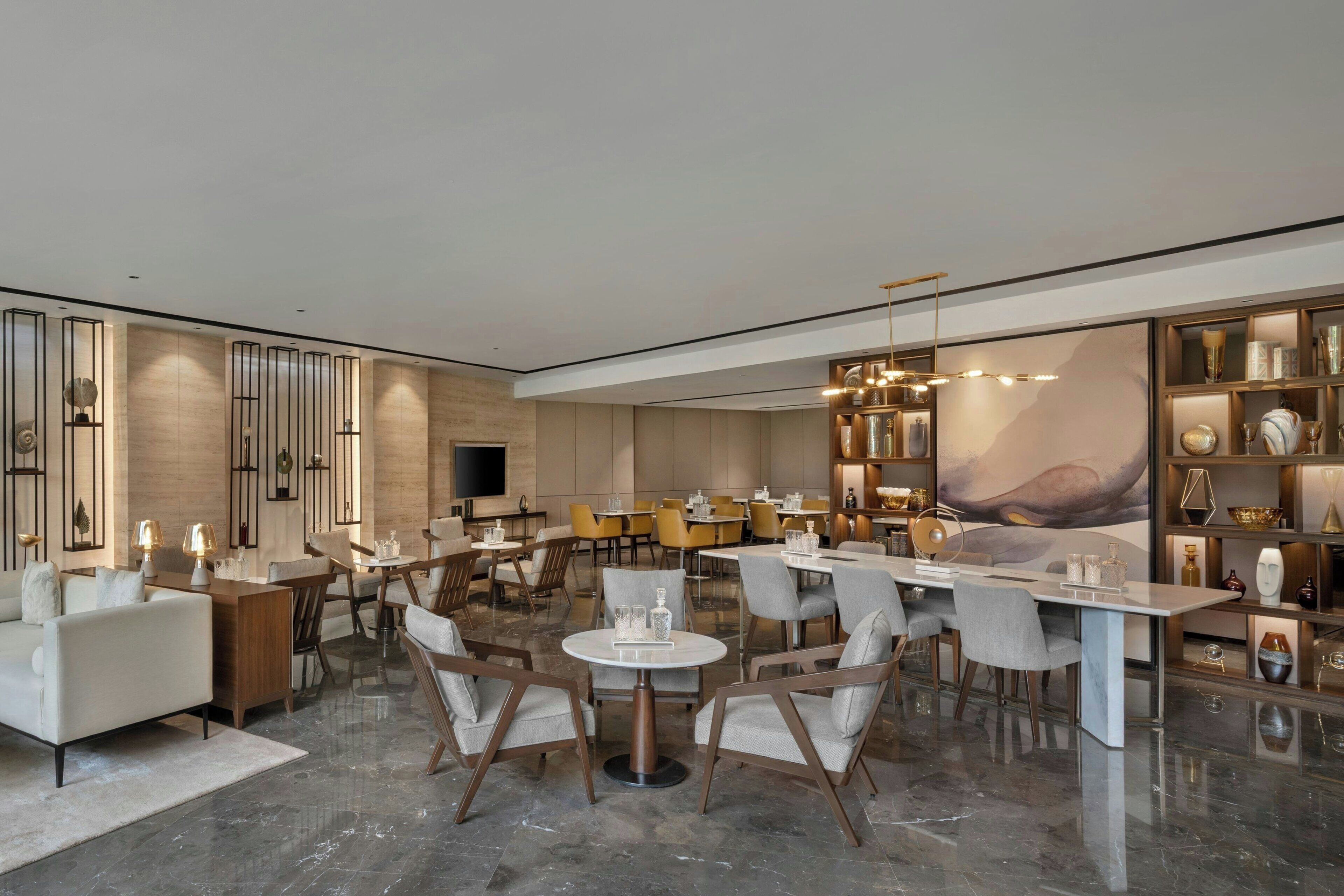 A modern hotel lobby with a mix of seating areas, including sofas and round tables, and a large bookshelf filled with decorative items.