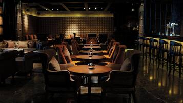 A dimly lit lounge area with leather sofas and round tables, a bar with stools, and a high ceiling with exposed beams.