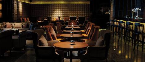 A dimly lit lounge area with leather sofas and round tables, a bar with stools, and a high ceiling with exposed beams.