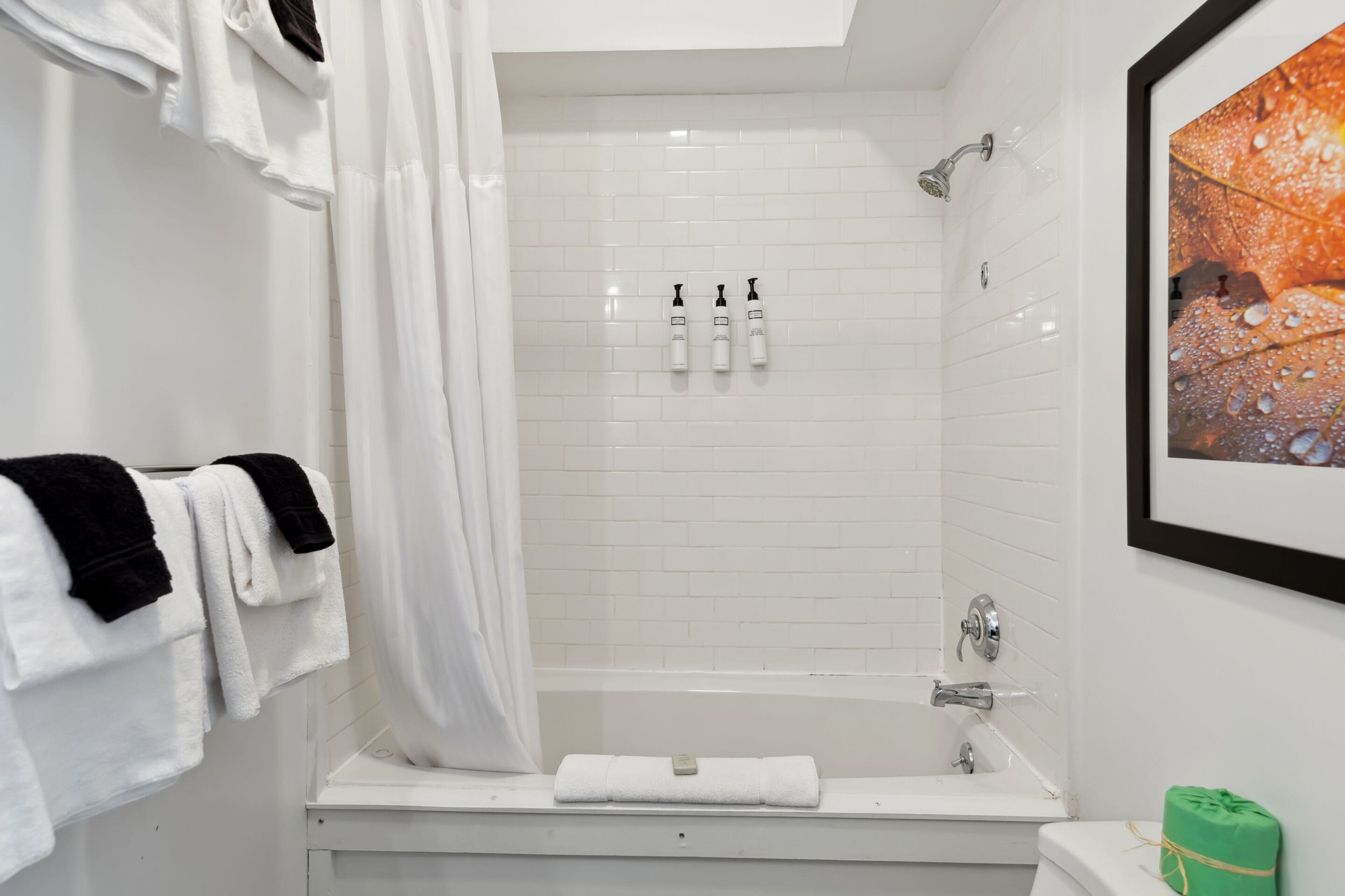 A modern bathroom with a white bathtub, a shower, and a toilet. There are towels hanging on a rack, and a framed picture on the wall.