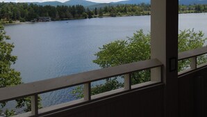 A view from a room with a wooden balcony overlooking a lake, surrounded by trees and mountains. - The Haus on Mirror Lake (Lake Placid)