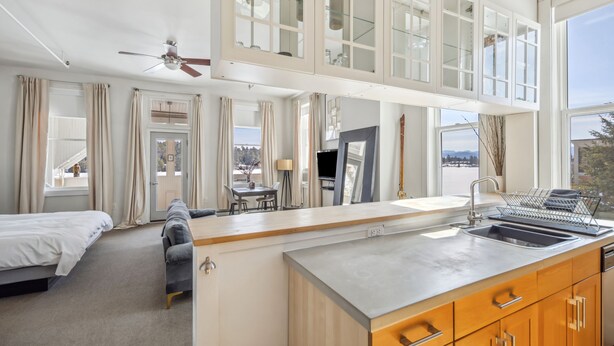 A modern hotel room with a large bed, a sofa, a dining area, a kitchen with a sink, and a balcony with a view. - The Haus on Mirror Lake (Lake Placid)