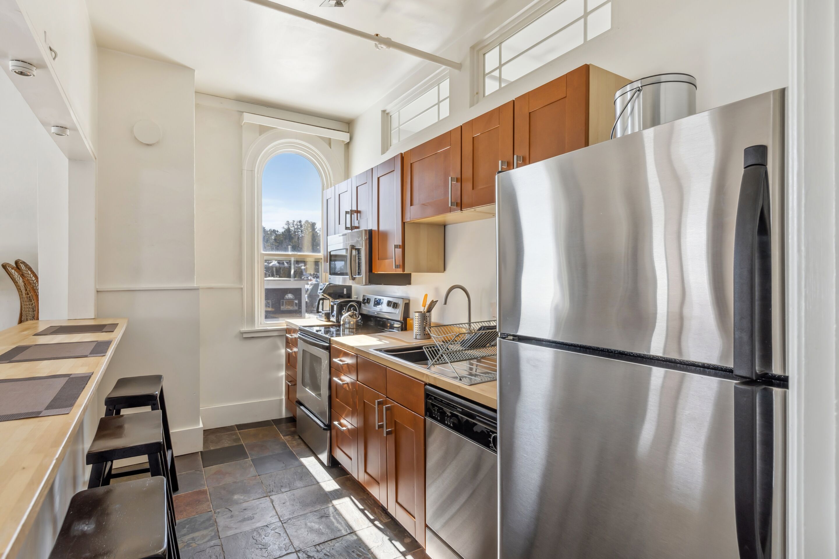 A modern kitchen with stainless steel appliances, wooden cabinets, and a large window providing natural light.