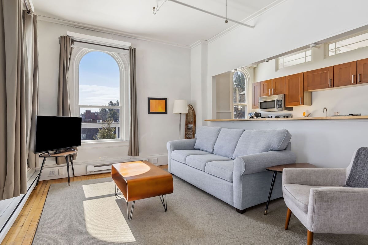 Un salon moderne avec un canapé gris, un pouf marron, un téléviseur posé sur un meuble et une vue sur la ville par une grande fenêtre.
