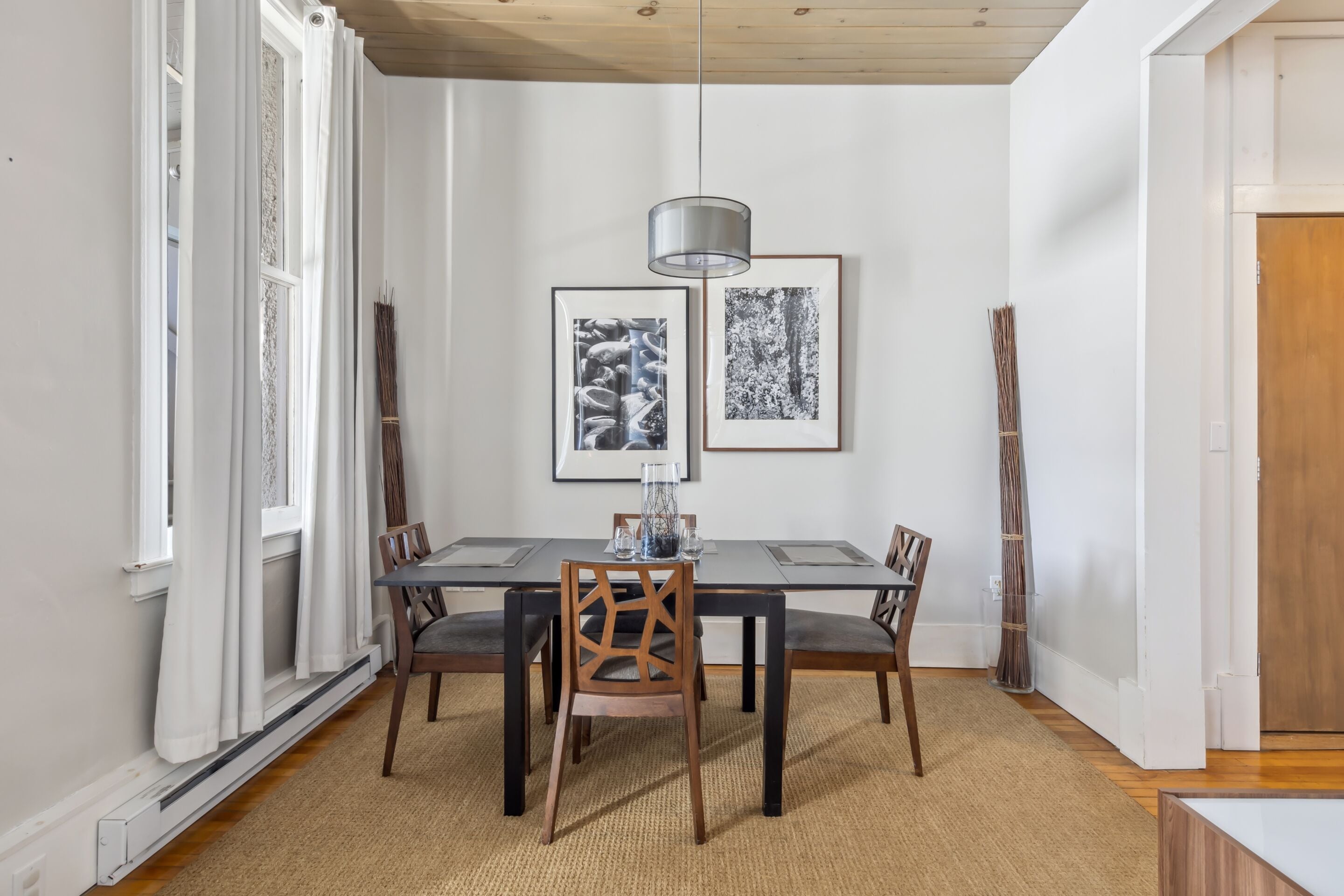 A dining area with a rectangular table, four chairs, a hanging pendant light, and two framed pictures on the wall.