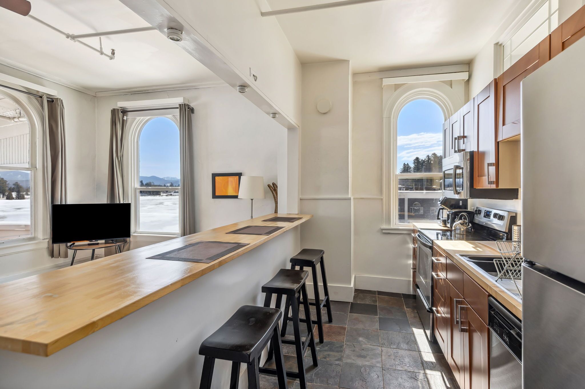 A modern kitchen with a long counter, bar stools, and a large window.
