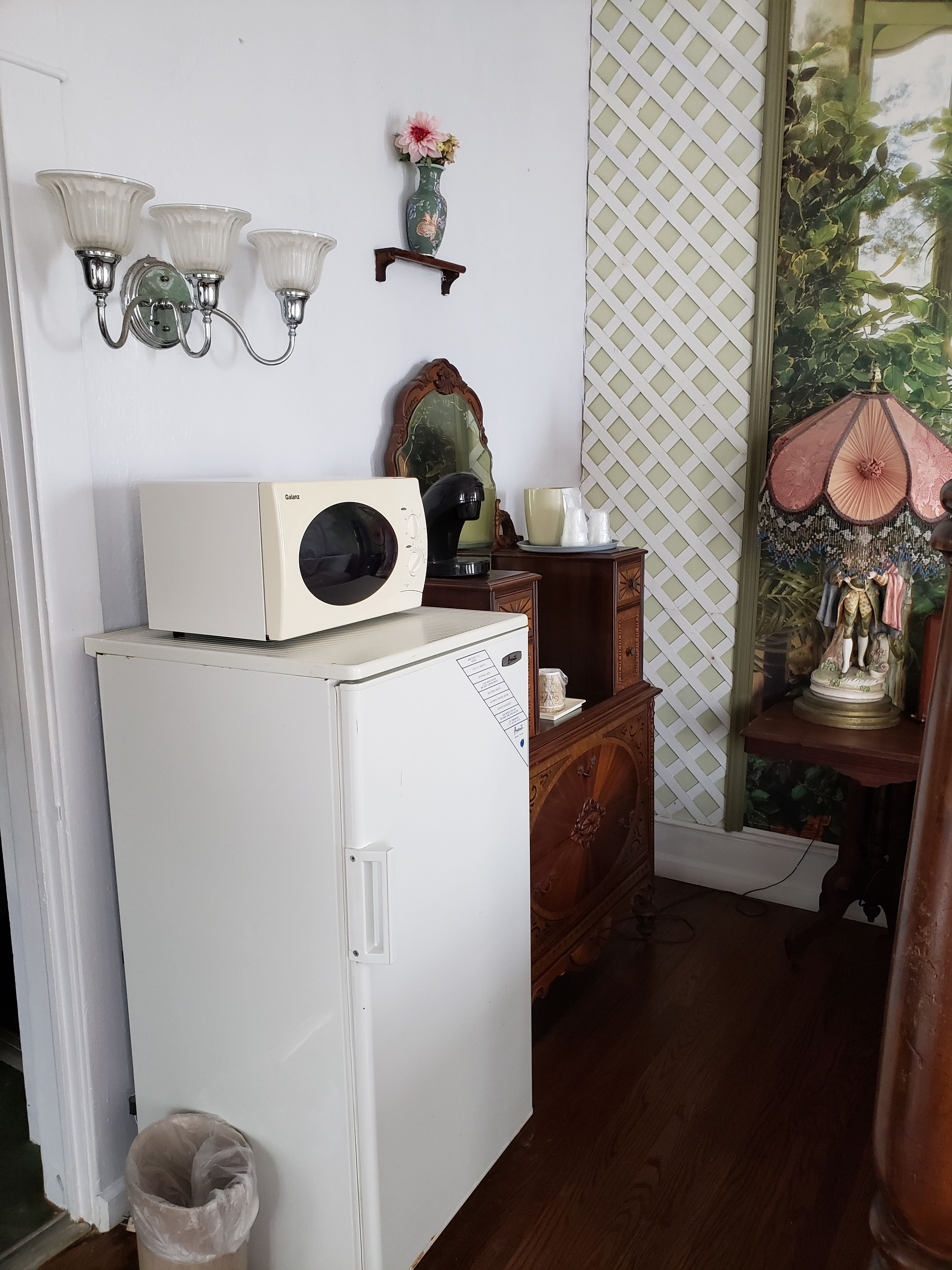 A compact room with a white refrigerator, a wooden dresser, a mirror, and a wall-mounted lamp.