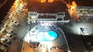 An aerial view of a hotel with a swimming pool surrounded by lounge chairs and umbrellas, multiple parking lots, and a large building with balconies. - Topaz Motel (Flagler Beach)