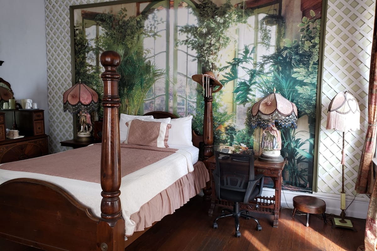 A bedroom with a four-poster bed, a desk with a chair, and a large tapestry depicting a tropical scene.