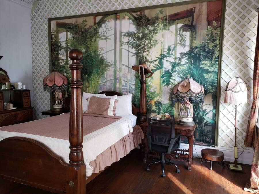A bedroom with a four-poster bed, a desk with a chair, and a large tapestry depicting a tropical scene.