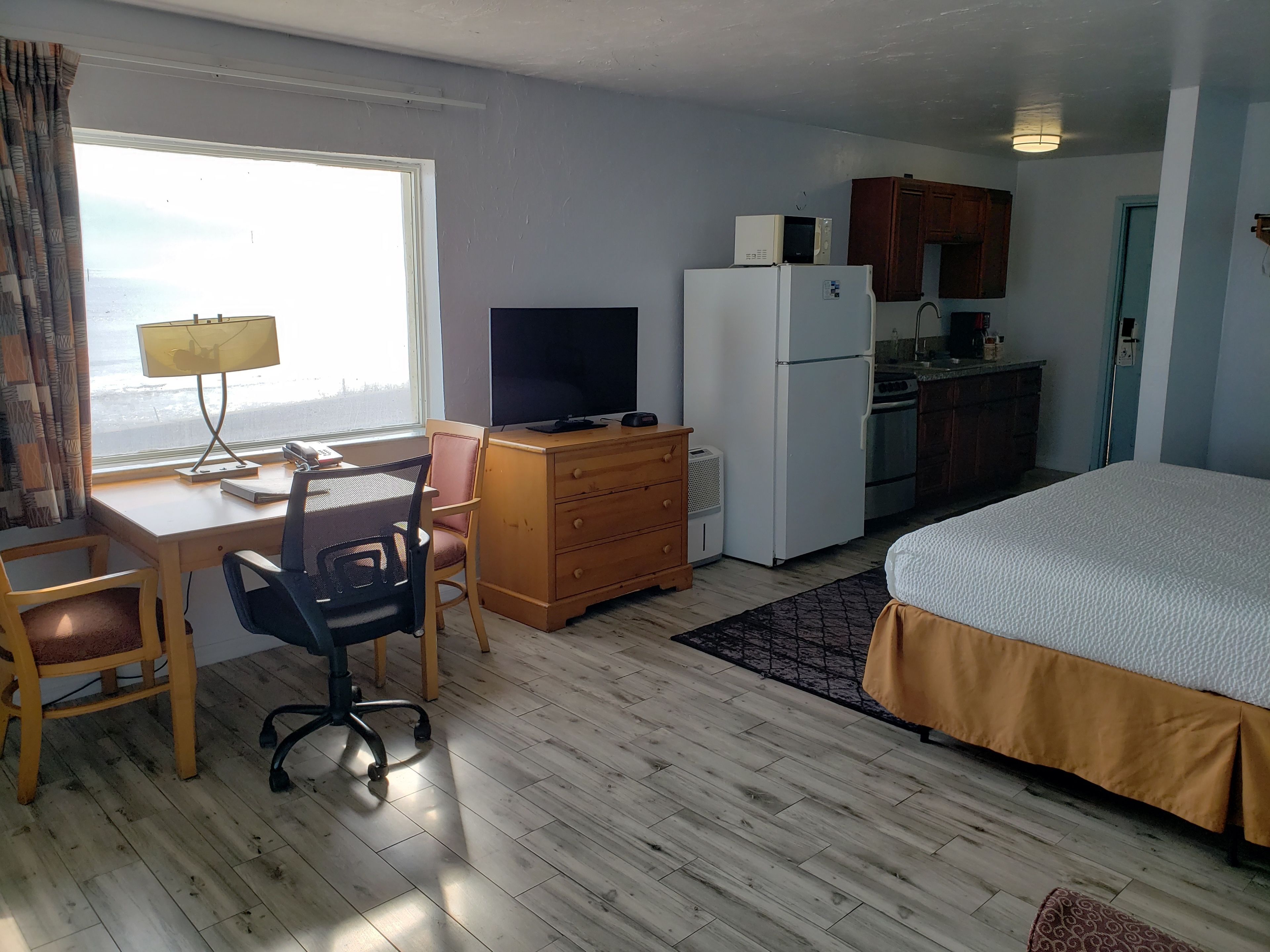 A hotel room with a bed, desk, chair, TV, refrigerator, and kitchenette.