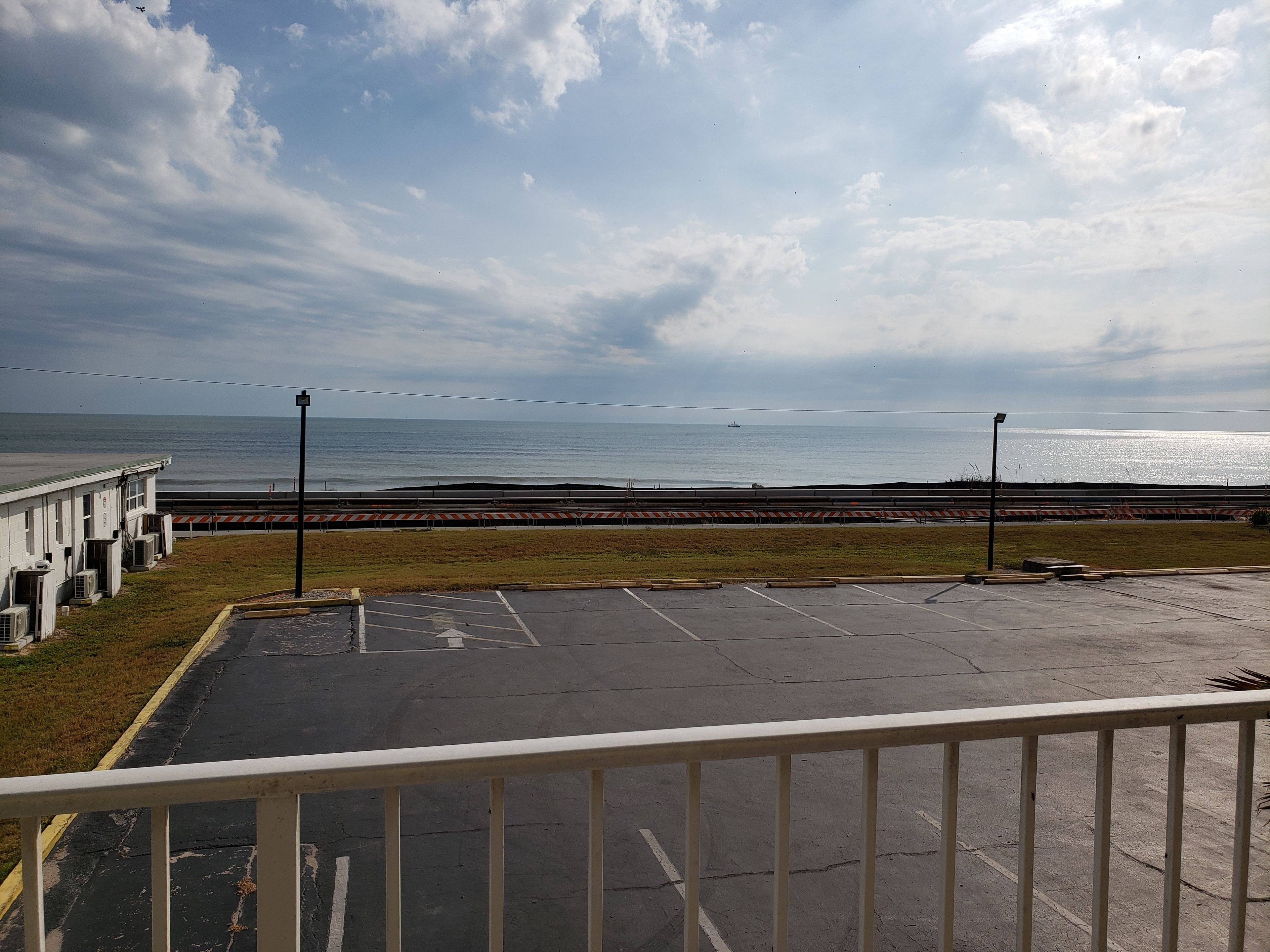 A coastal view with a parking area, a building with air conditioning units, and a distant ship on the water.