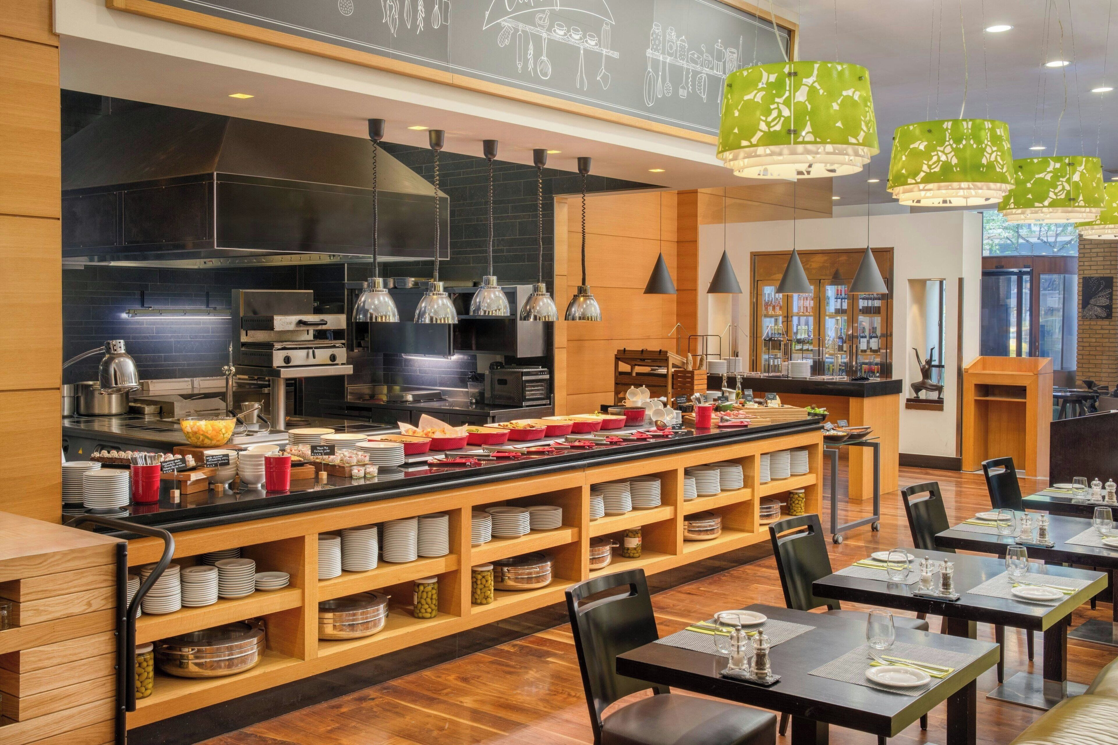 A modern restaurant with a central buffet area, multiple dining tables, and a kitchen in the background.