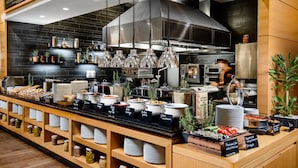 Un área de bufé con distintas estaciones de comida, que incluyen barra de ensaladas, estación de carnes y estación de postres. Se observan también varios recipientes y utensilios para servir.