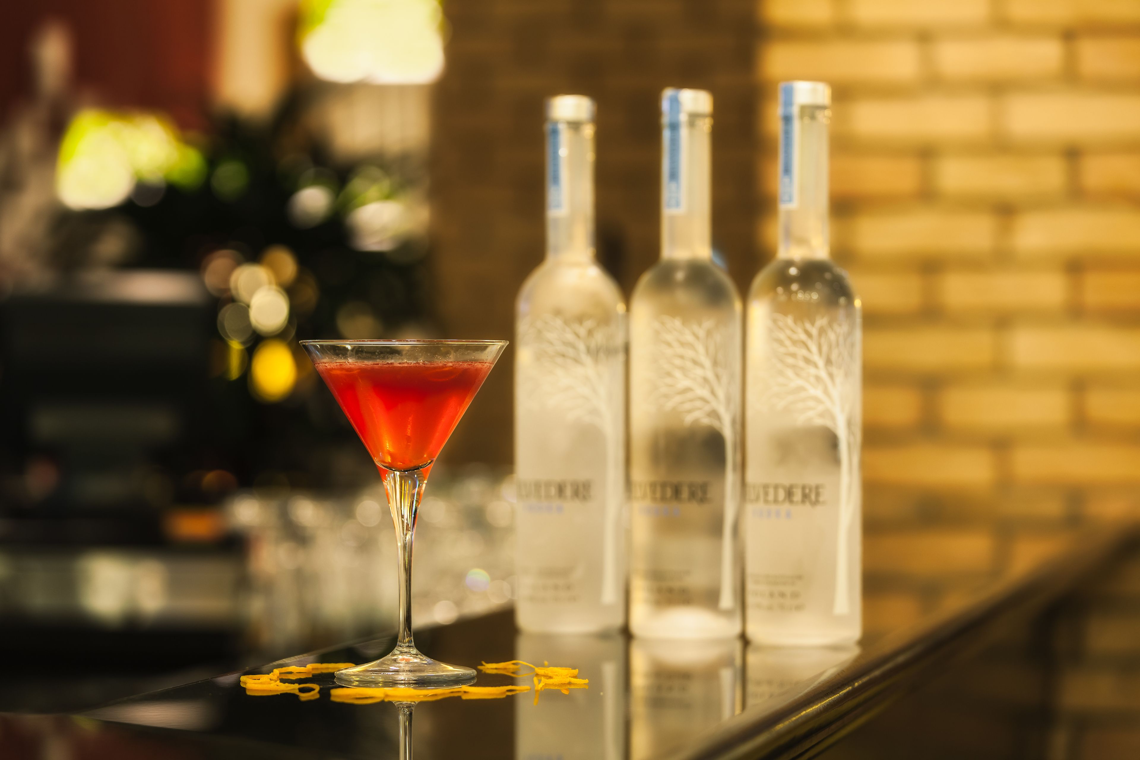 Un verre à martini contenant une boisson rouge, posé sur le comptoir d’un bar, avec trois bouteilles de spiritueux derrière.