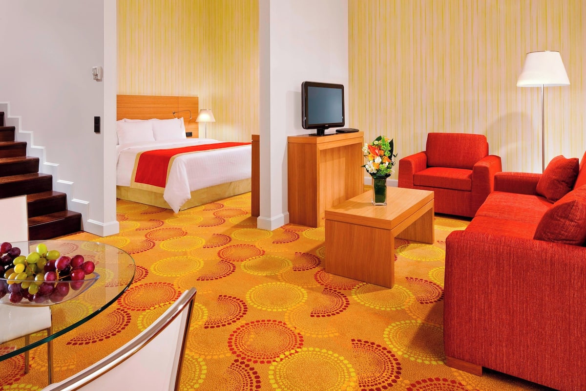 A hotel room with a bed, a TV, a red sofa, a glass table with fruit, and a carpet with circular patterns.