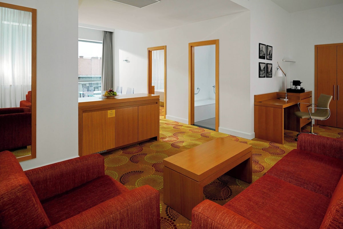 A modern hotel room with a wooden desk, a red sofa, a small table, and a view of the city through large windows.