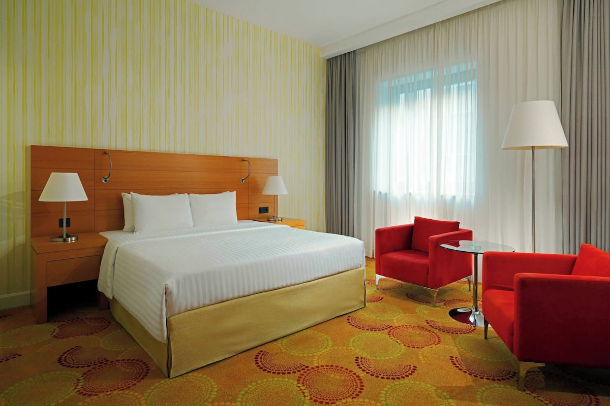 A hotel room with a large bed, two bedside tables with lamps, a red armchair, a small round table, and a large window with curtains.