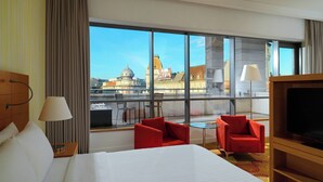 Habitación de hotel con una cama grande, dos sillas rojas, una mesita, un televisor y un amplio ventanal con vista a la ciudad.