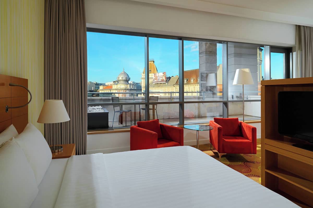 A hotel room with a large bed, two red chairs, a small table, a TV, and a large window with a city view.