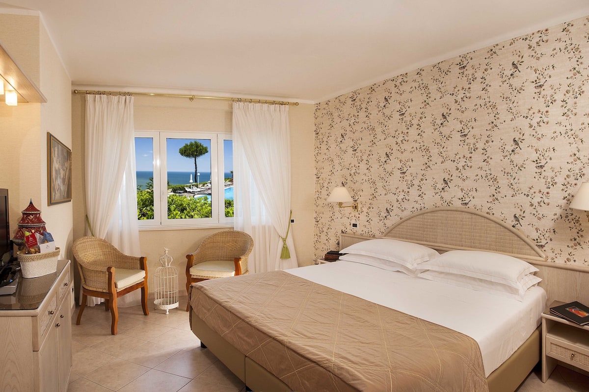 A hotel room with a bed, two chairs, a desk, a TV, and a window with a view of the sea.