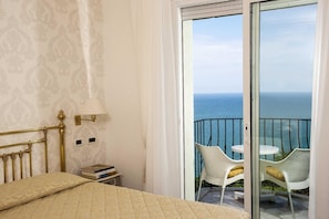 A bedroom with a bed, a nightstand, a lamp, and a view of the sea through a balcony door. - Posillipo (Gabicce Mare)
