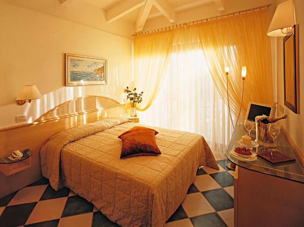 Habitación de hotel con cama, mesita de noche, lámpara, ventana con cortinas y una pequeña mesa con una botella y vasos.
