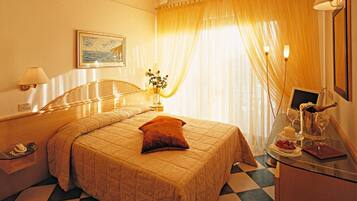 Habitación de hotel con cama, mesita de noche, lámpara, ventana con cortinas y una pequeña mesa con una botella y vasos.