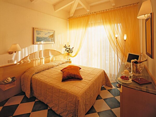Quarto de hotel com cama, mesa de cabeceira, luminária, janela com cortinas e uma pequena mesa com garrafa e copos.