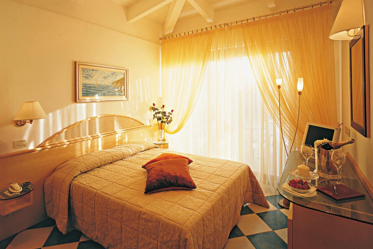 A hotel room with a bed, a nightstand, a table with a bottle and glasses, and a painting on the wall.
